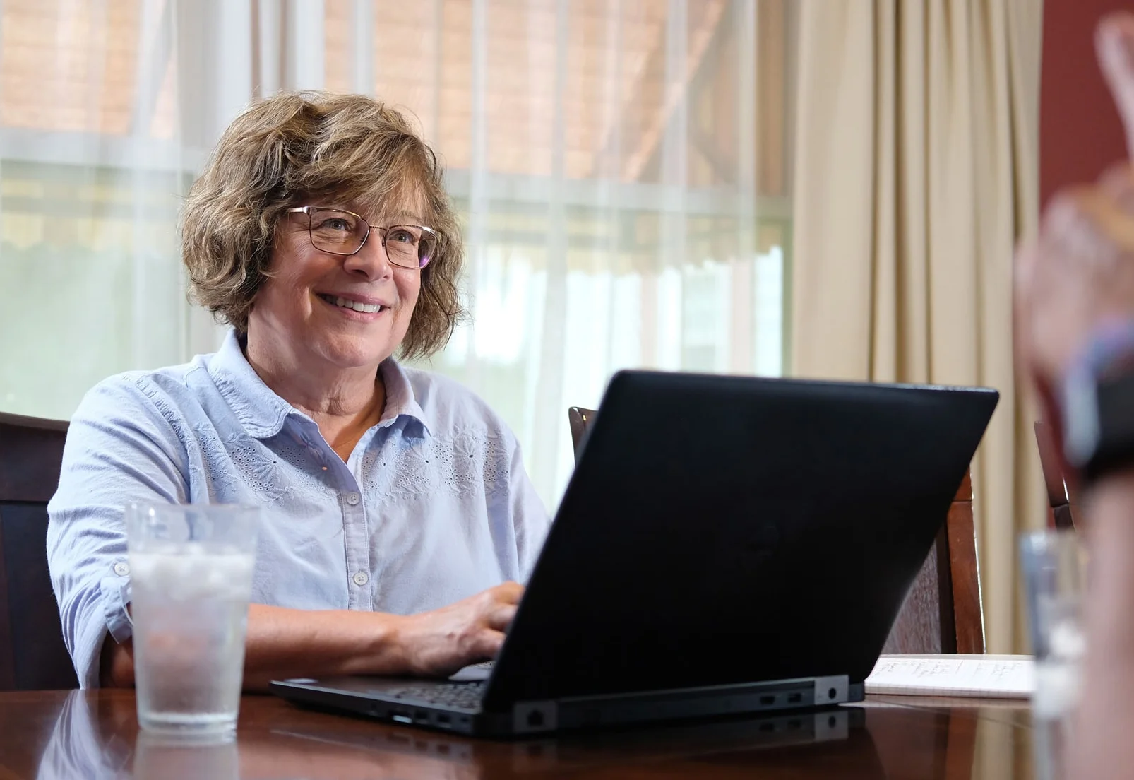 A smiling woman with glasses sitting at a table, using a laptop, with a glass of water nearby.