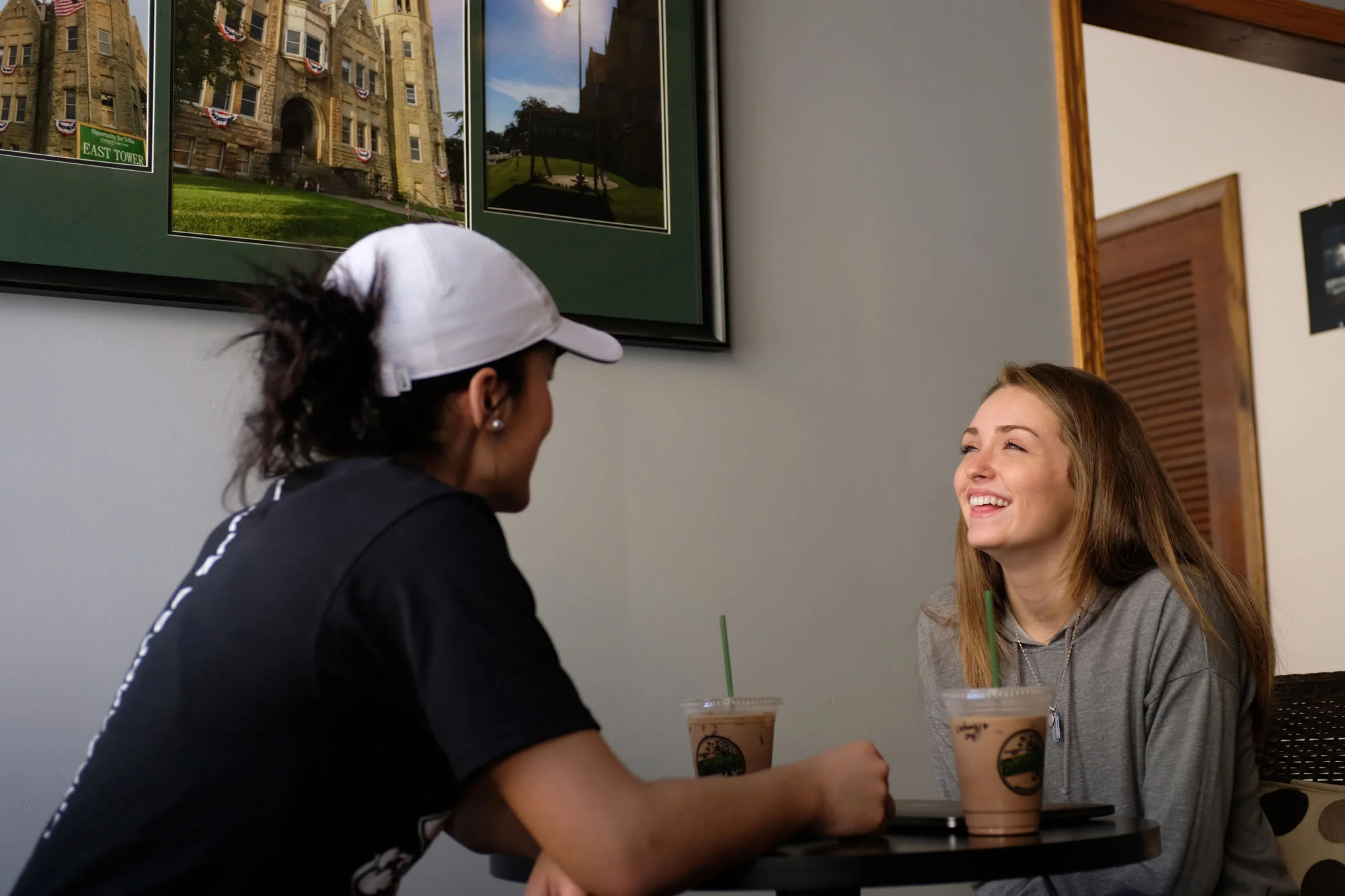 Two women sitting at a table, smiling and talking, with iced drinks in front of them, inside a cafe or restaurant. One woman wears a white cap and black shirt, the other has long blonde hair and a gray hoodie.