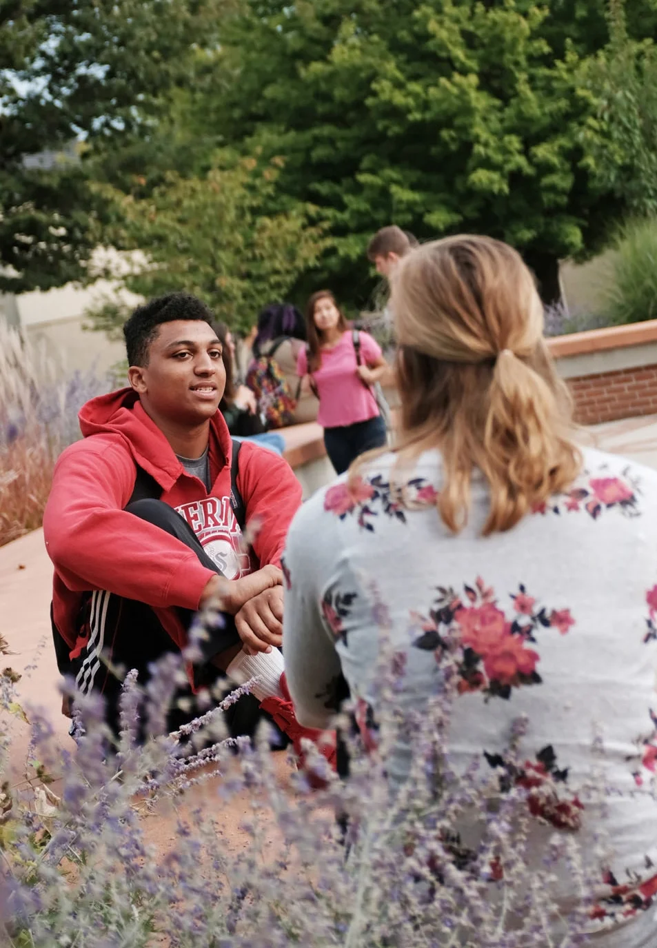 A young man sitting on the ground and talking to a woman with light brown hair tied back, outdoors in a park with trees and purple flowers, while several other people walk in the background.
