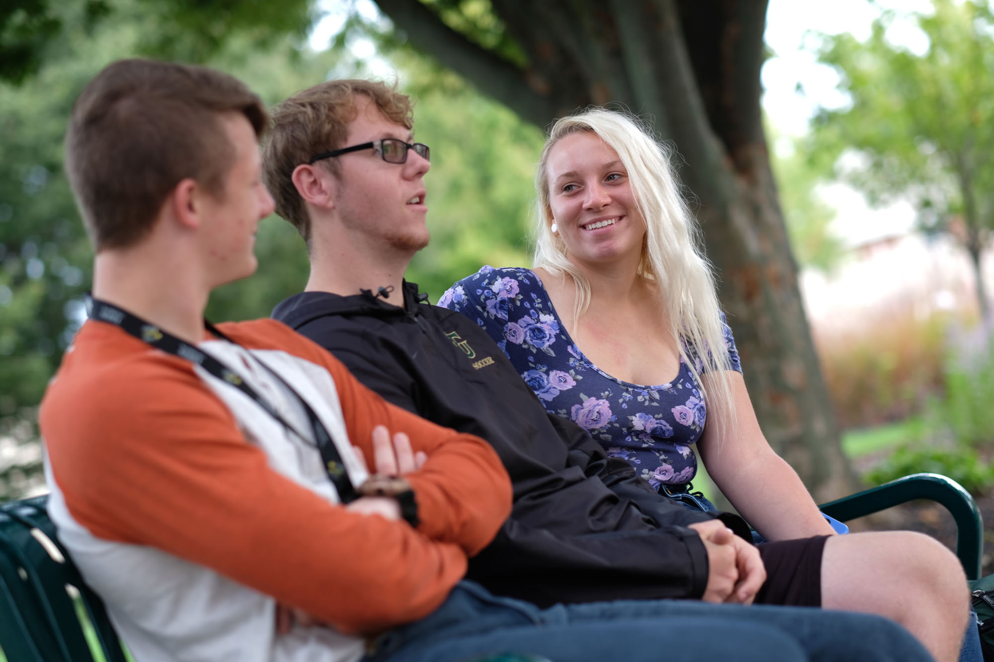 Three young adults sitting on a park bench under trees, engaged in conversation.