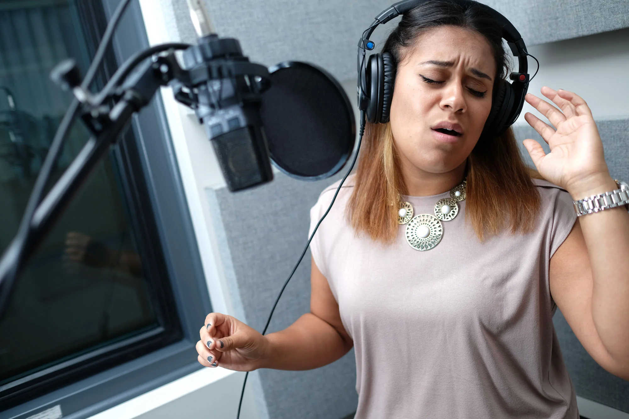 A woman singing into a microphone at a recording studio, wearing large over-ear headphones and a statement necklace.