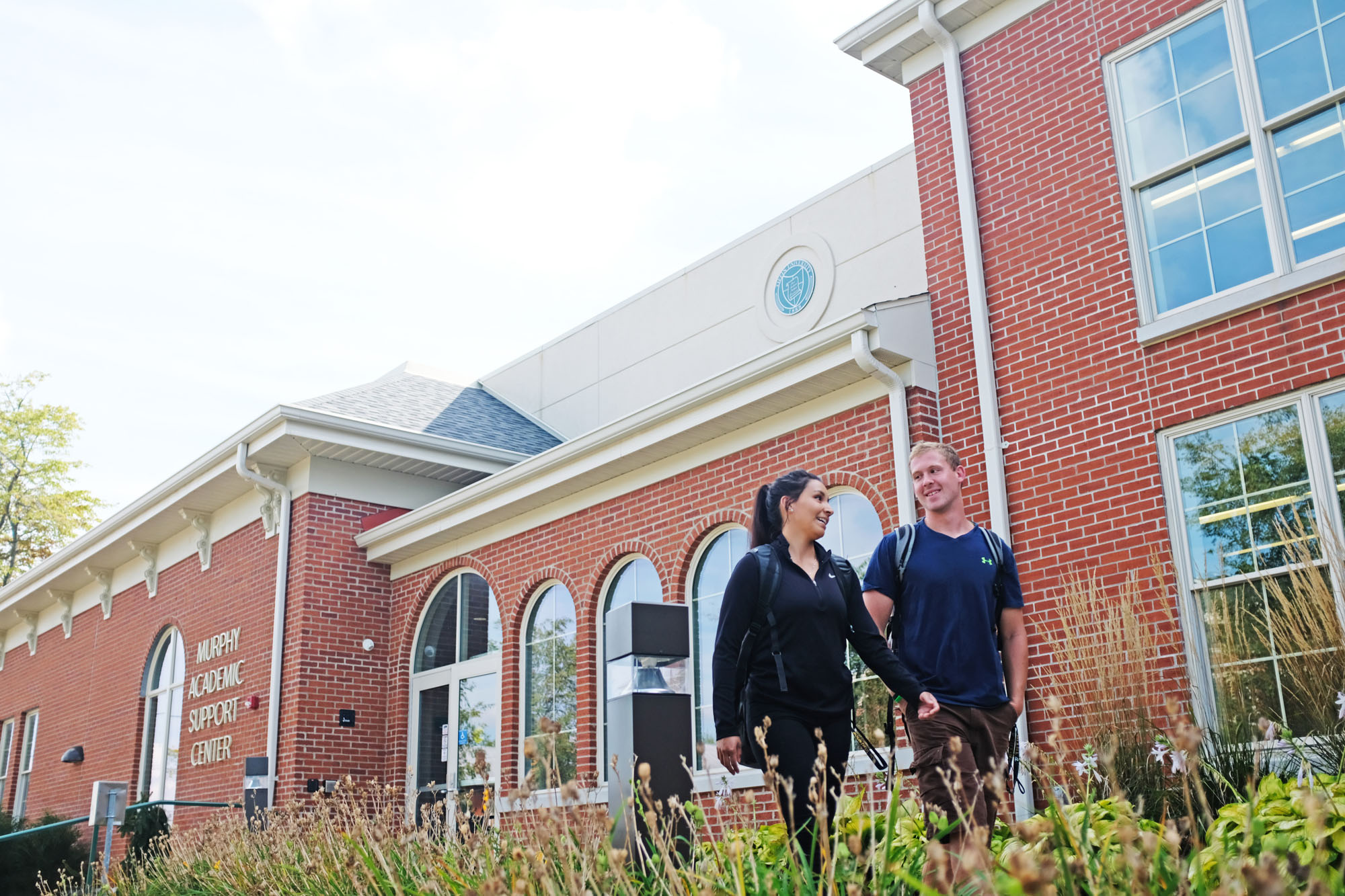 Two students walking and talking outside the Murphy Academic Support Center, a red brick building with large windows.