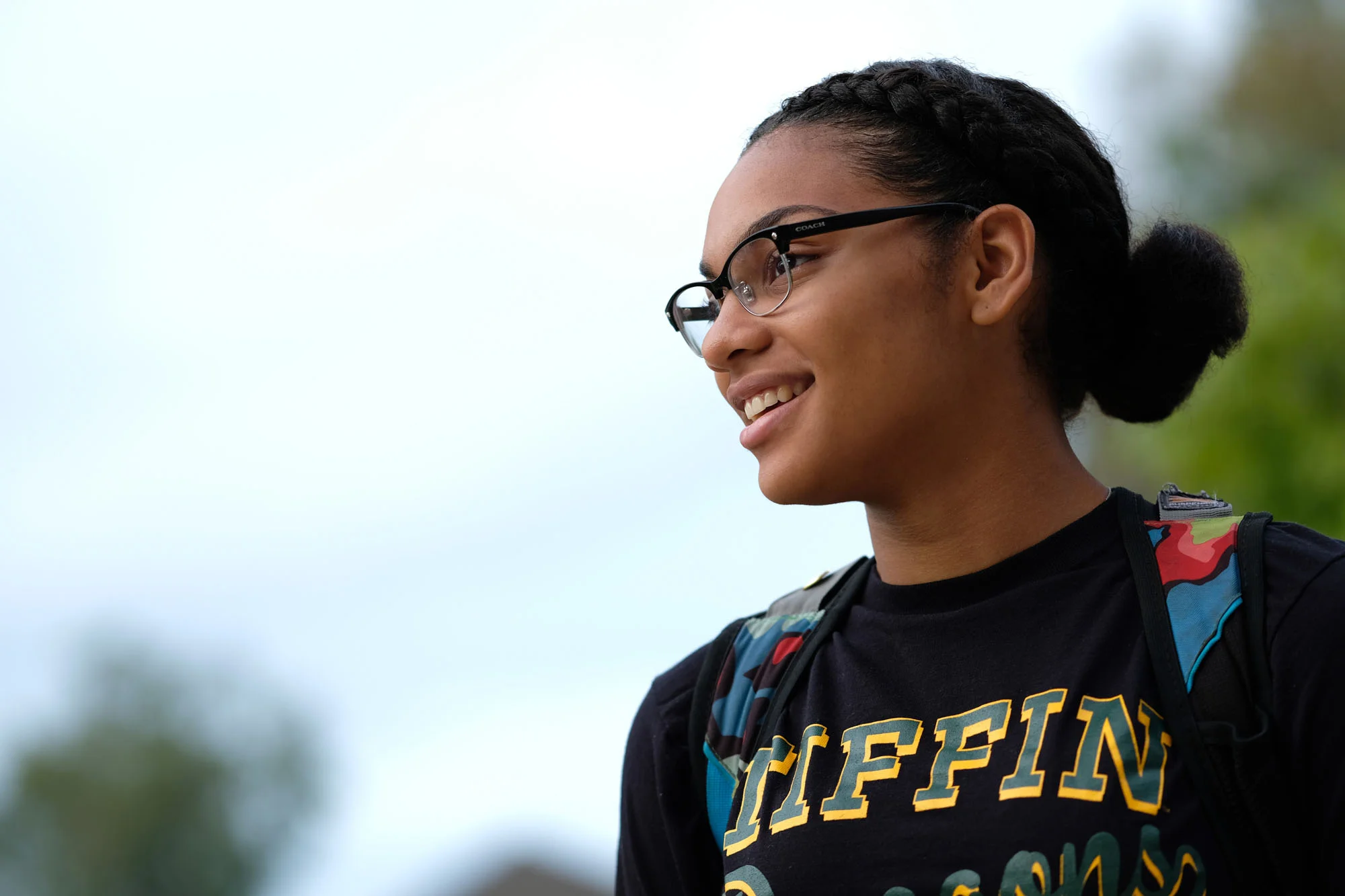 A young woman with glasses and braided hair smiling outdoors, wearing a black T-shirt with yellow and green text and a colorful backpack.