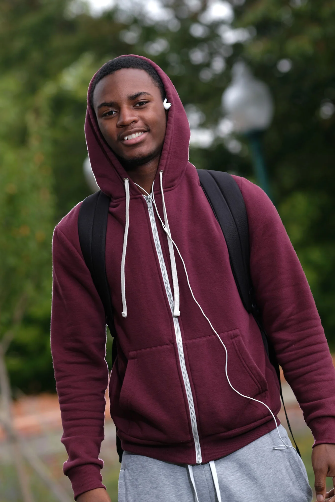 A young man wearing a maroon hoodie with the hood up, gray sweatpants, and a black backpack. He has earbuds in his ears and is standing outdoors with green trees and a lamp post in the background.