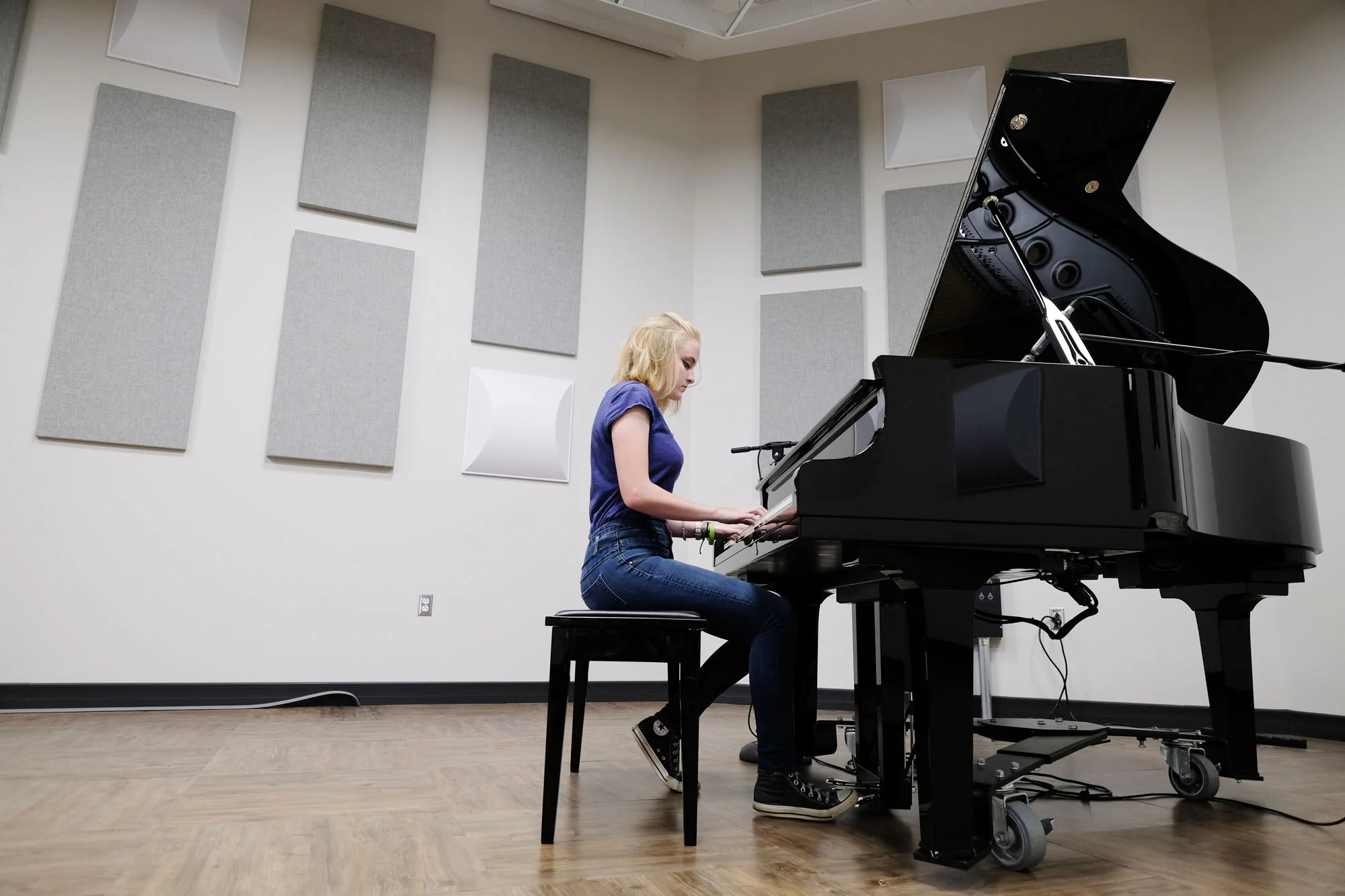 A young woman with blonde hair wearing a blue t-shirt and jeans sitting on a stool playing a grand piano in a music studio.