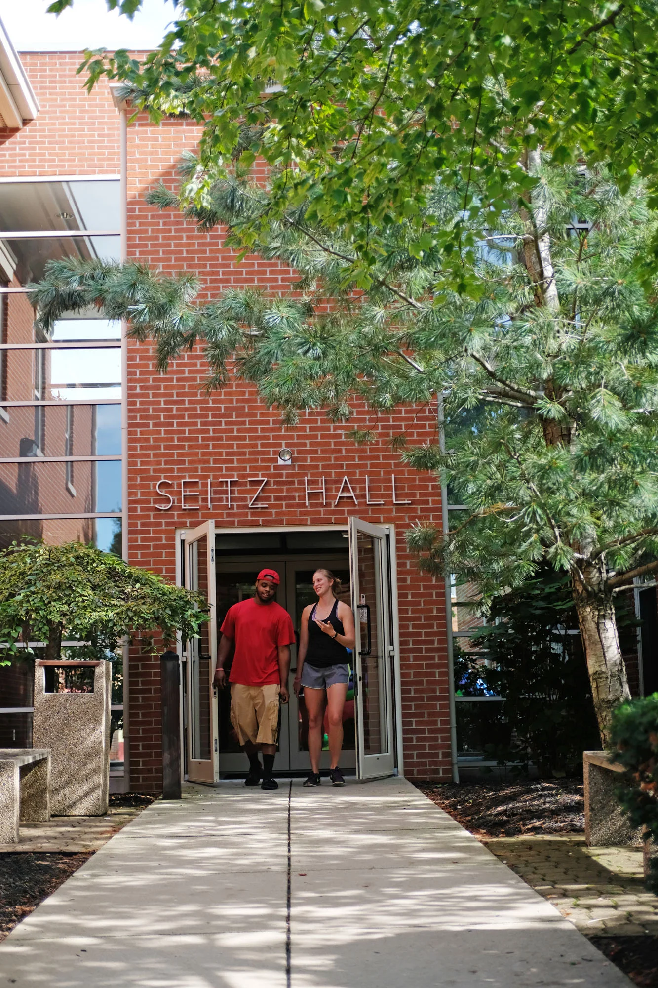Two students, a young man in a red T-shirt and khaki shorts and a young woman in a black tank top and gray shorts, walk out of Seitz Hall entrance on a college campus during daytime, surrounded by trees and bushes.