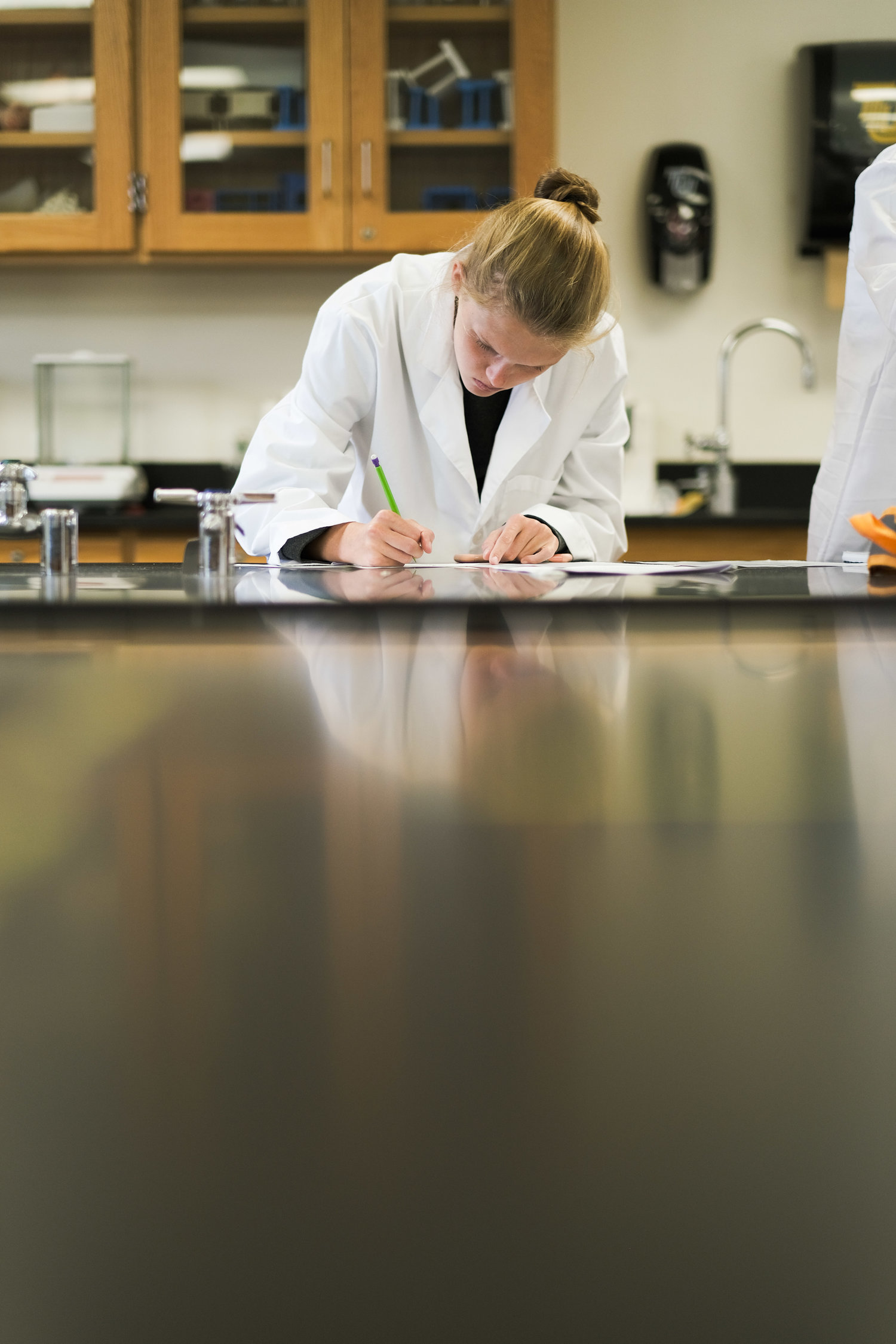 A girl in a lab coat writing on a sheet in a science classroom or laboratory.