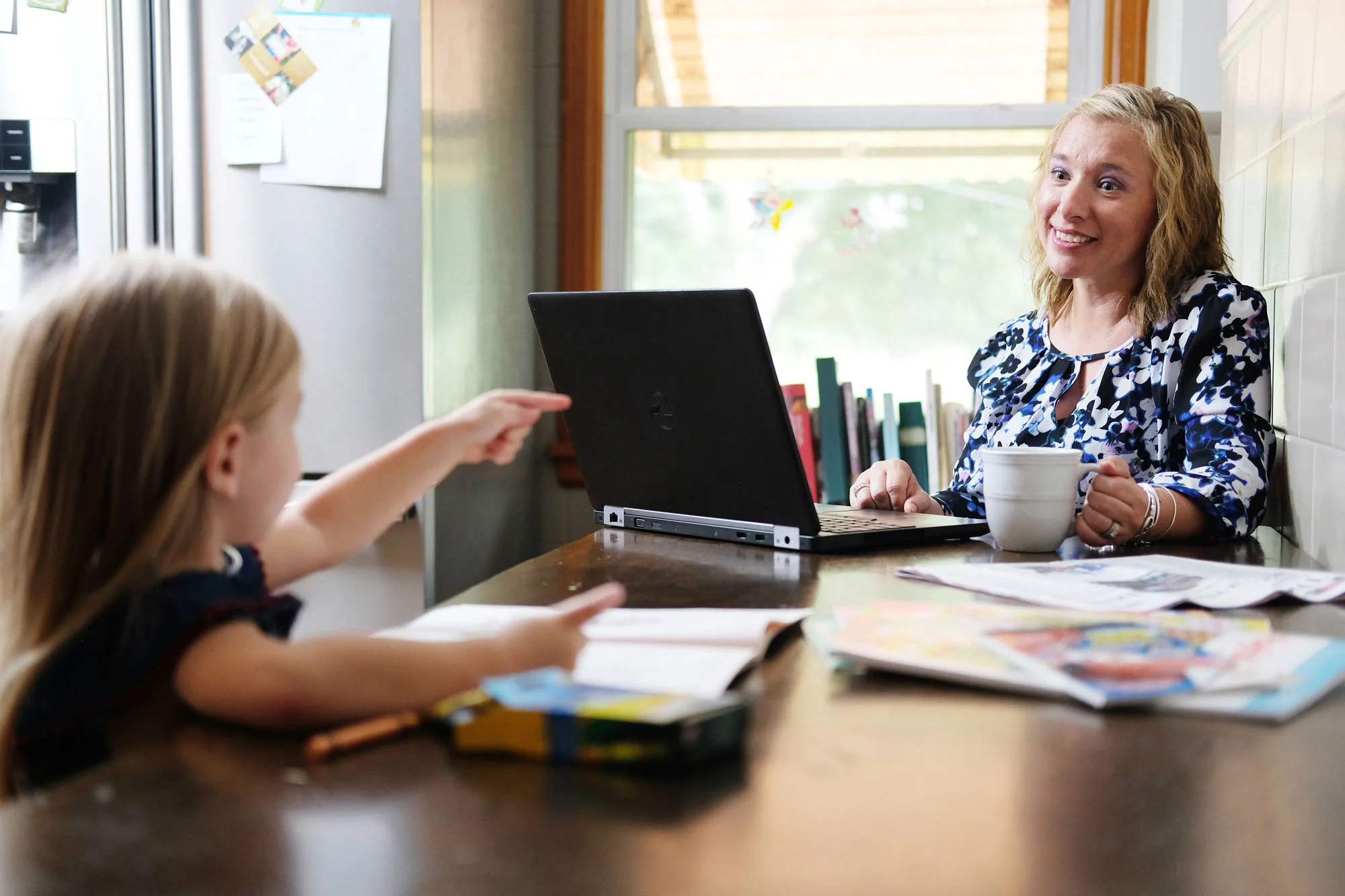 A woman and a young girl sitting at a kitchen table, engaging in a conversation. The woman is smiling and holding a mug, while the girl points at her with her finger. There are books and papers on the table, and a coffee mug is also visible.