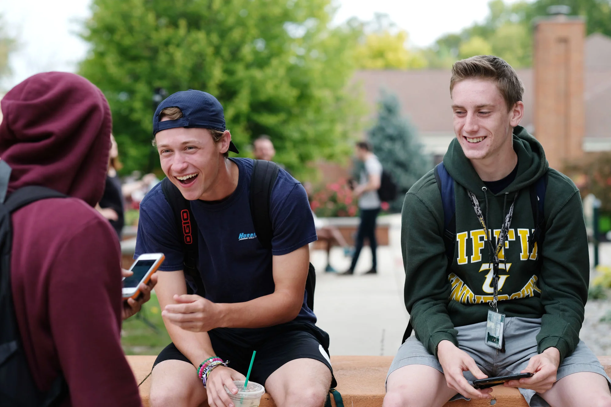 Two young men sitting on a bench outdoors, smiling and talking to a person wearing a maroon hoodie holding a phone.