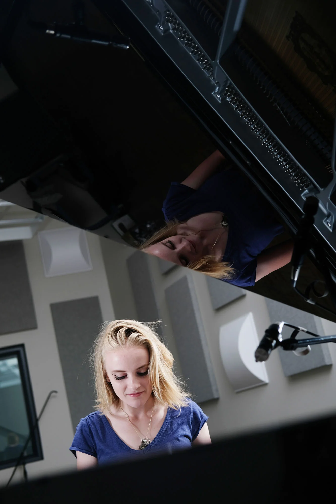 A young woman with blonde hair wearing a blue shirt is seen from below, looking down at a piano with an open lid, in a music studio with soundproofing panels on the walls.