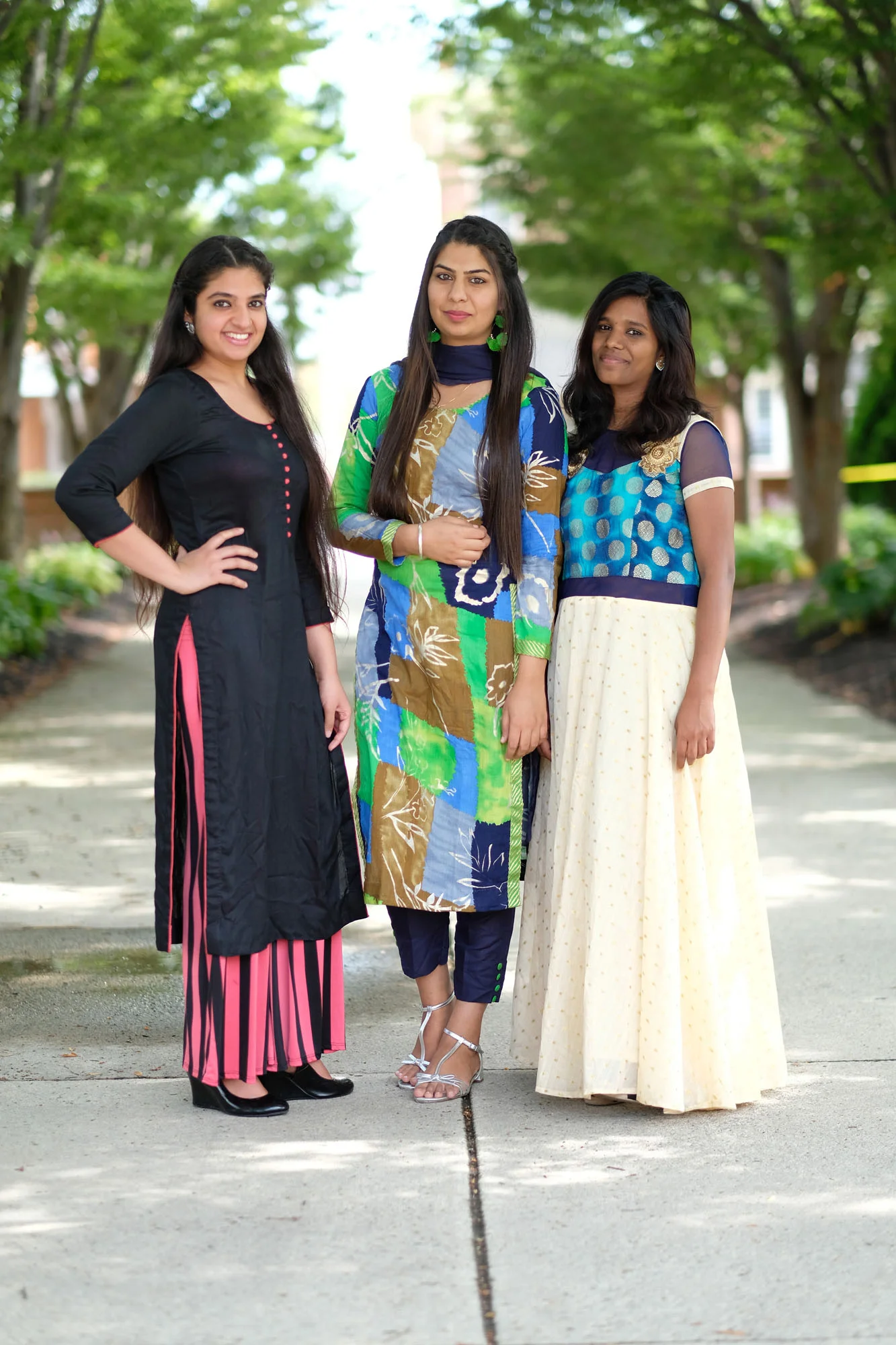 Three young women standing outdoors on a sidewalk, wearing colorful traditional and modern clothing, smiling and posing for the camera with trees and a building in the background.