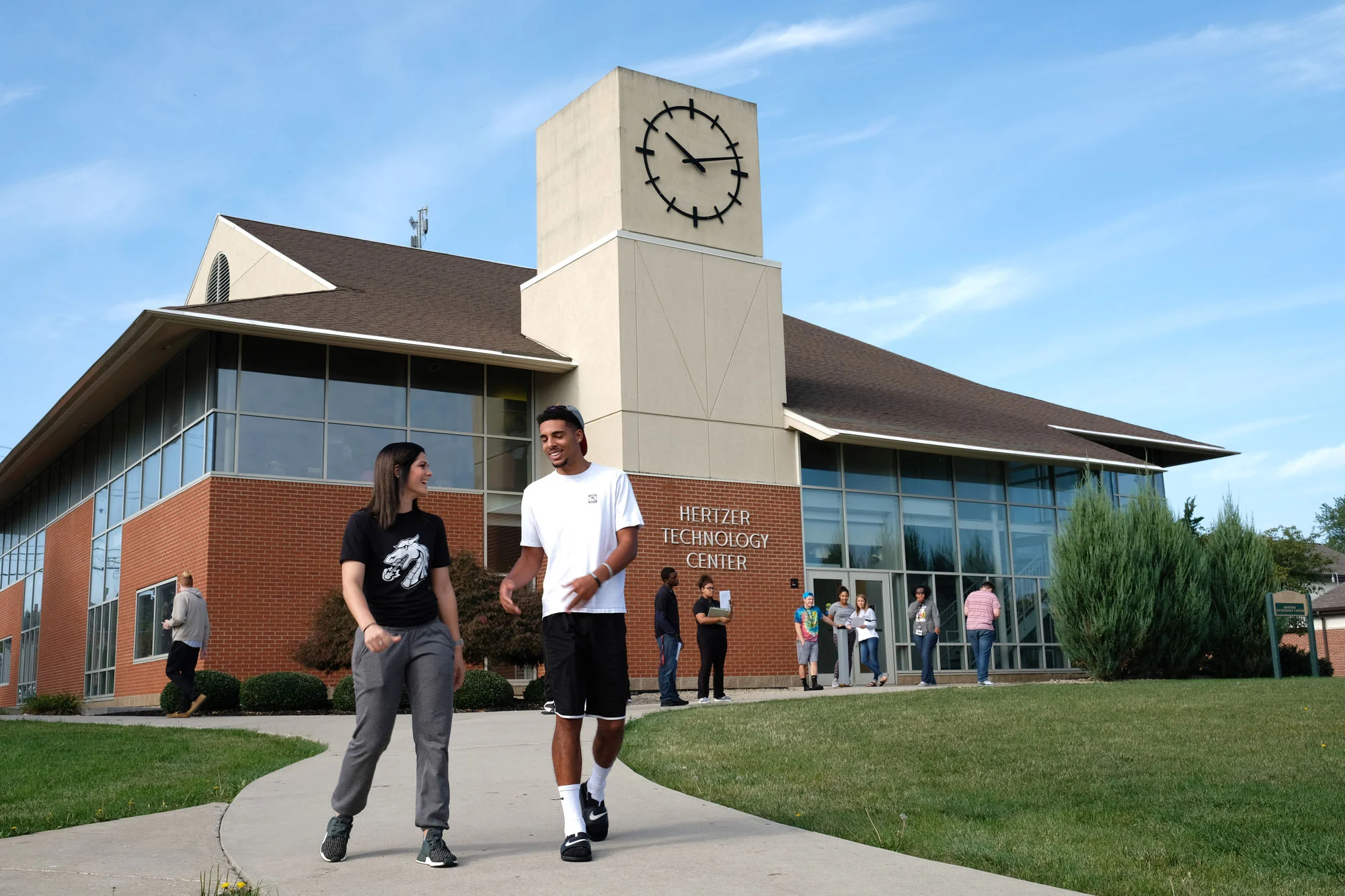 Students walking and chatting outside the Hertzzer Technology Center, a modern building with large glass windows and a brick facade, under a clear blue sky.