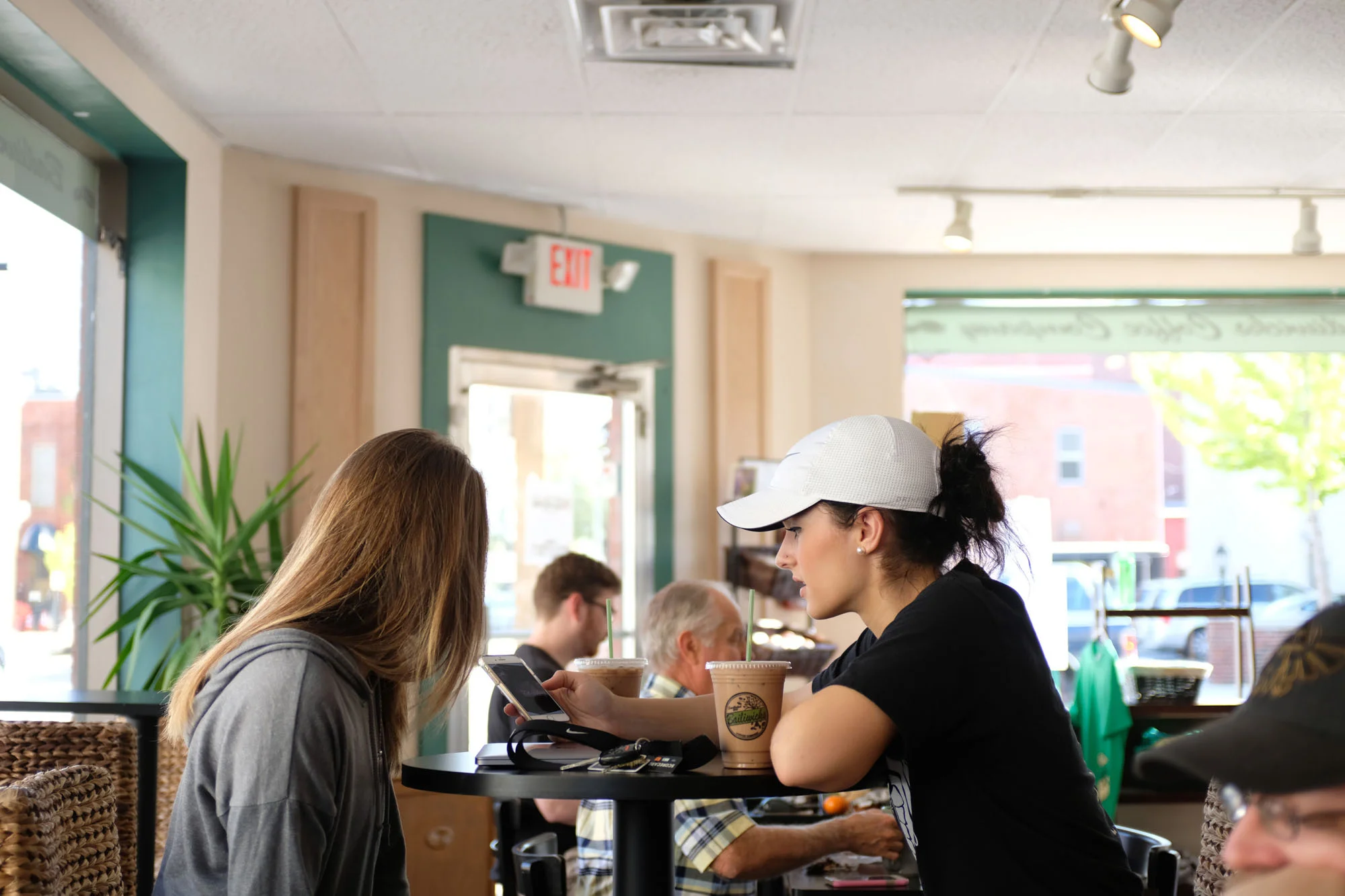 Two women sit at a table in a cafe, one with long hair and a gray hoodie, the other with a white cap, black shirt, and dark hair in a bun, looking at her phone. There are iced drinks on the table.