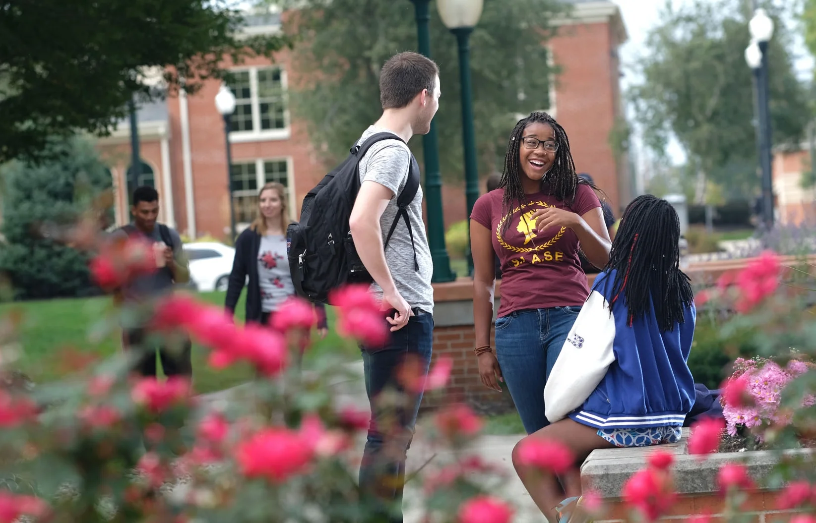 Four college students outdoors on campus, with pink flowers in the foreground. One girl seated on a bench talking to a standing girl, while two others are walking and looking at their phones.