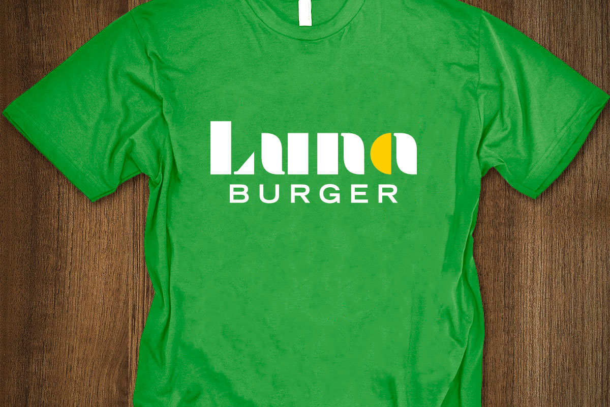 Green T-shirt with a logo that says 'Luna BURGER' in stylized white and yellow text, laid flat on a wooden surface.