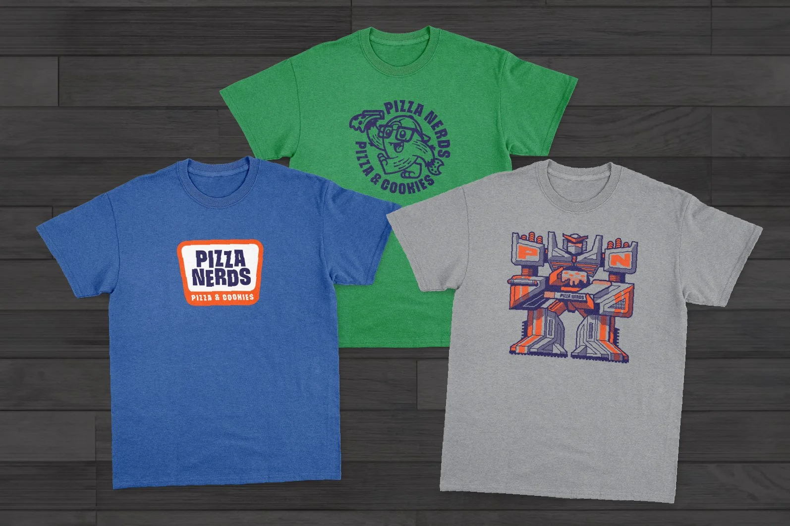 Three T-shirts laid flat on a dark wooden surface. The T-shirt on the left is blue with a pizza and cookies logo that says "Pizza Nerds." The T-shirt in the middle is green with a cartoon pizza character holding a remote, surrounded by the text "Pizz