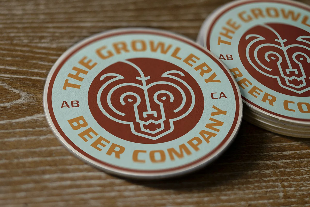 Round sticker for The Growler Beer Company featuring a stylized bear face in red with orange and light blue text, placed on a wooden surface.