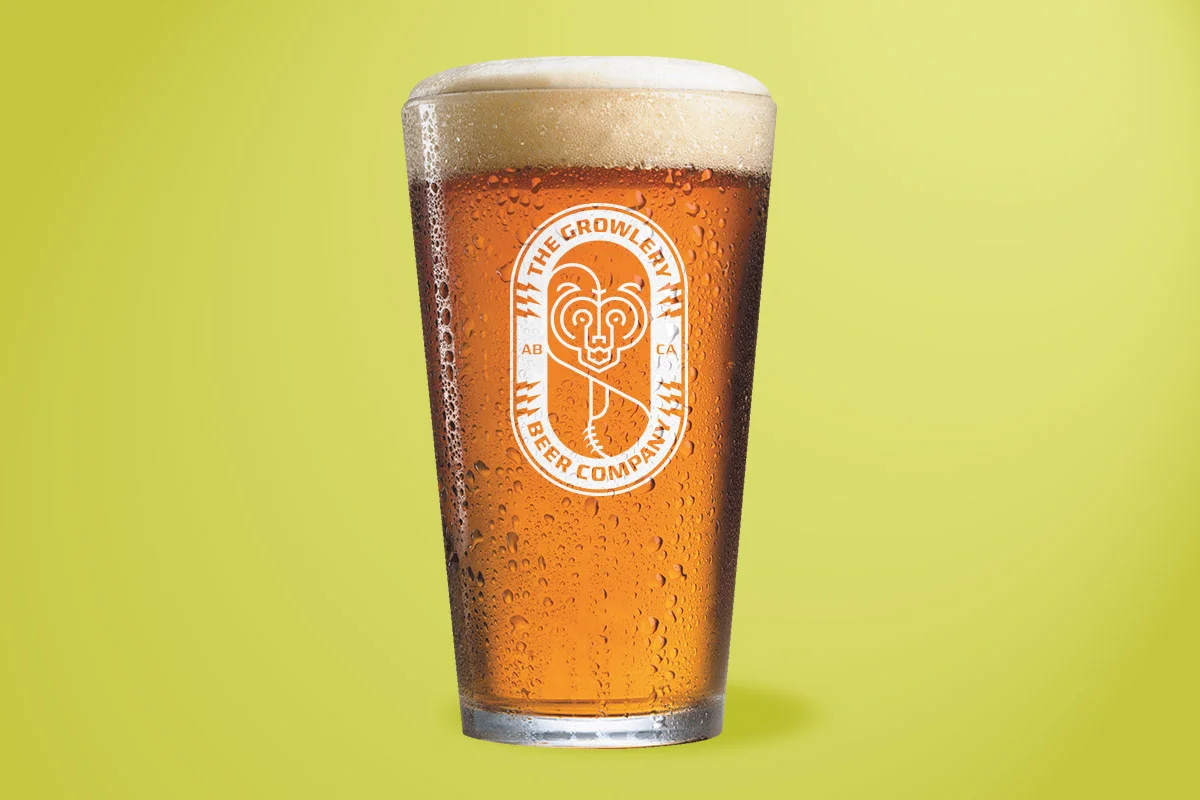 A pint glass filled with amber beer, condensation on the outside, and a foamy head on top, with a logo of a lion's face in an oval and the text 'The Growlery Beer Company' on the glass, against a gradient yellow-green background.