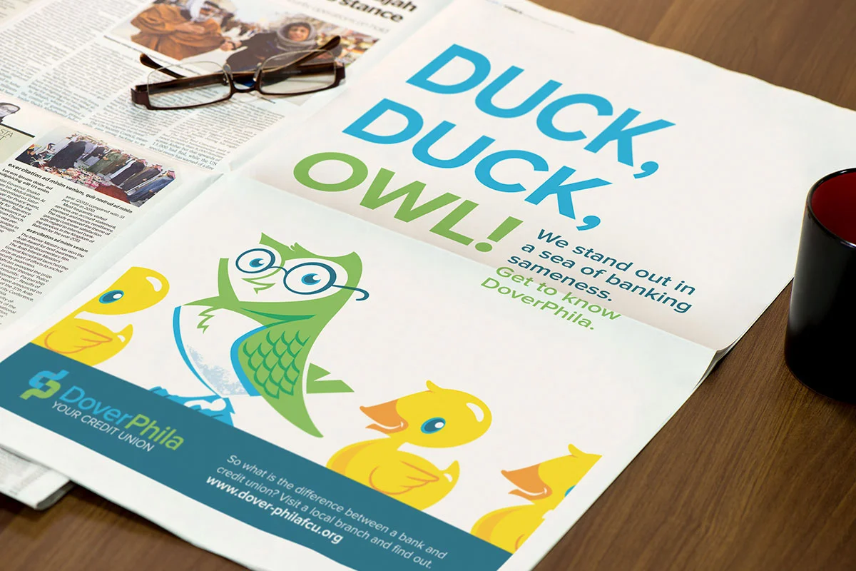 A colorful poster promoting Dover Federal Credit Union with the slogan "Duck, Duck, OWL!" and cartoon characters of an owl and three ducks. The poster includes text about banking and a website link, with a pair of glasses resting on a newspaper on a 