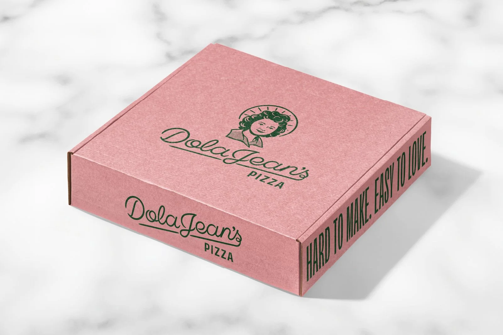 Pink pizza box from DolaJean's Pizza with a logo of a woman's face and the text 'DolaJean's Pizza' and a slogan 'HARD TO MAKE. EASY TO LOVE.' on a white marble surface.