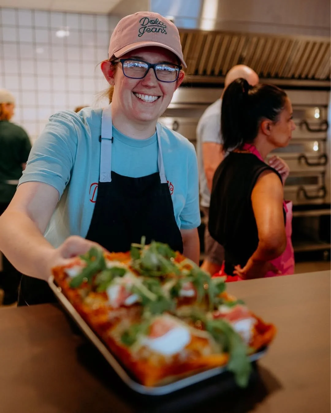 A smiling woman wearing glasses, a beige cap with 'Deli Jean's' embroidered on it, a light blue t-shirt, and a black apron, holding a tray of food in a busy kitchen or restaurant setting with other staff members working in the background.