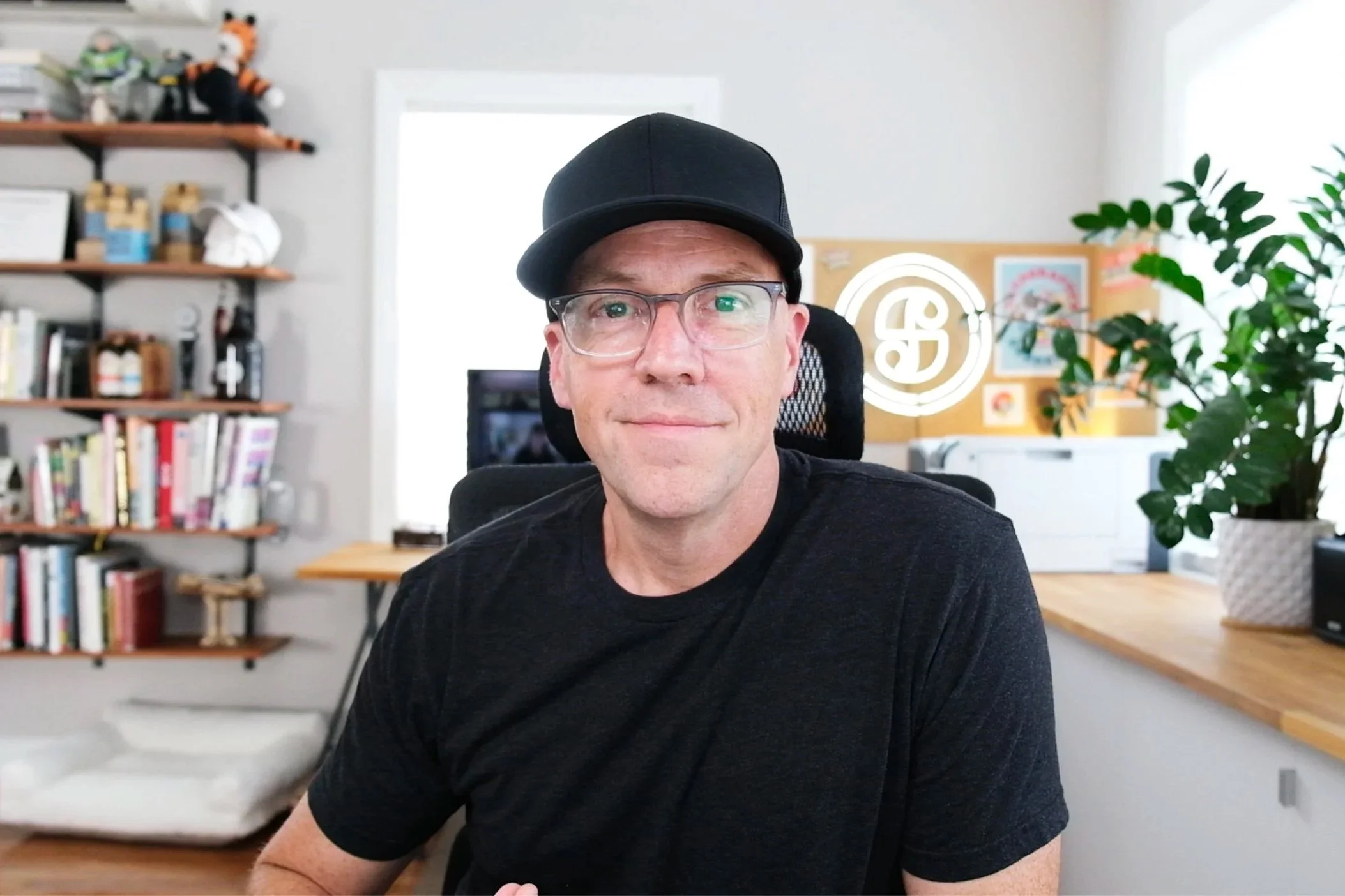 A man wearing a black baseball cap, glasses, and a black t-shirt sitting at a desk in a well-lit home office, with bookshelves, a neon sign, and houseplants in the background.