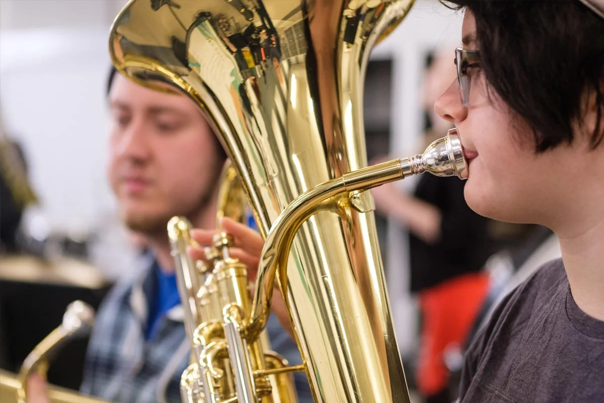 Two young people playing brass instruments, a tuba and a saxophone, in a music rehearsal or band setting.