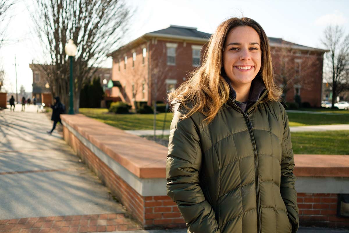 A young woman smiling outdoors on a sunny day, wearing a green puffer jacket, with a brick building and trees in the background.
