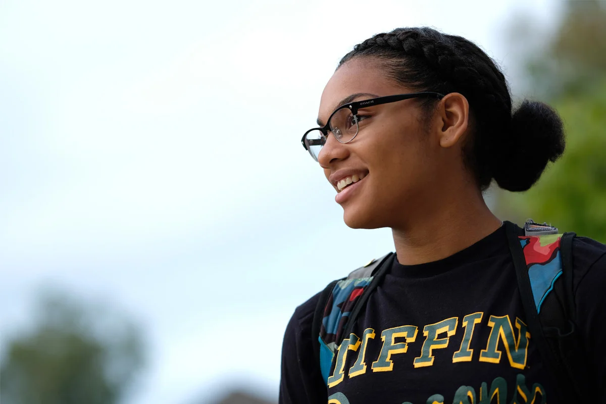 Profile of a smiling young woman with glasses, wearing a black T-shirt and a backpack, outdoors with a blurred background of trees and sky.