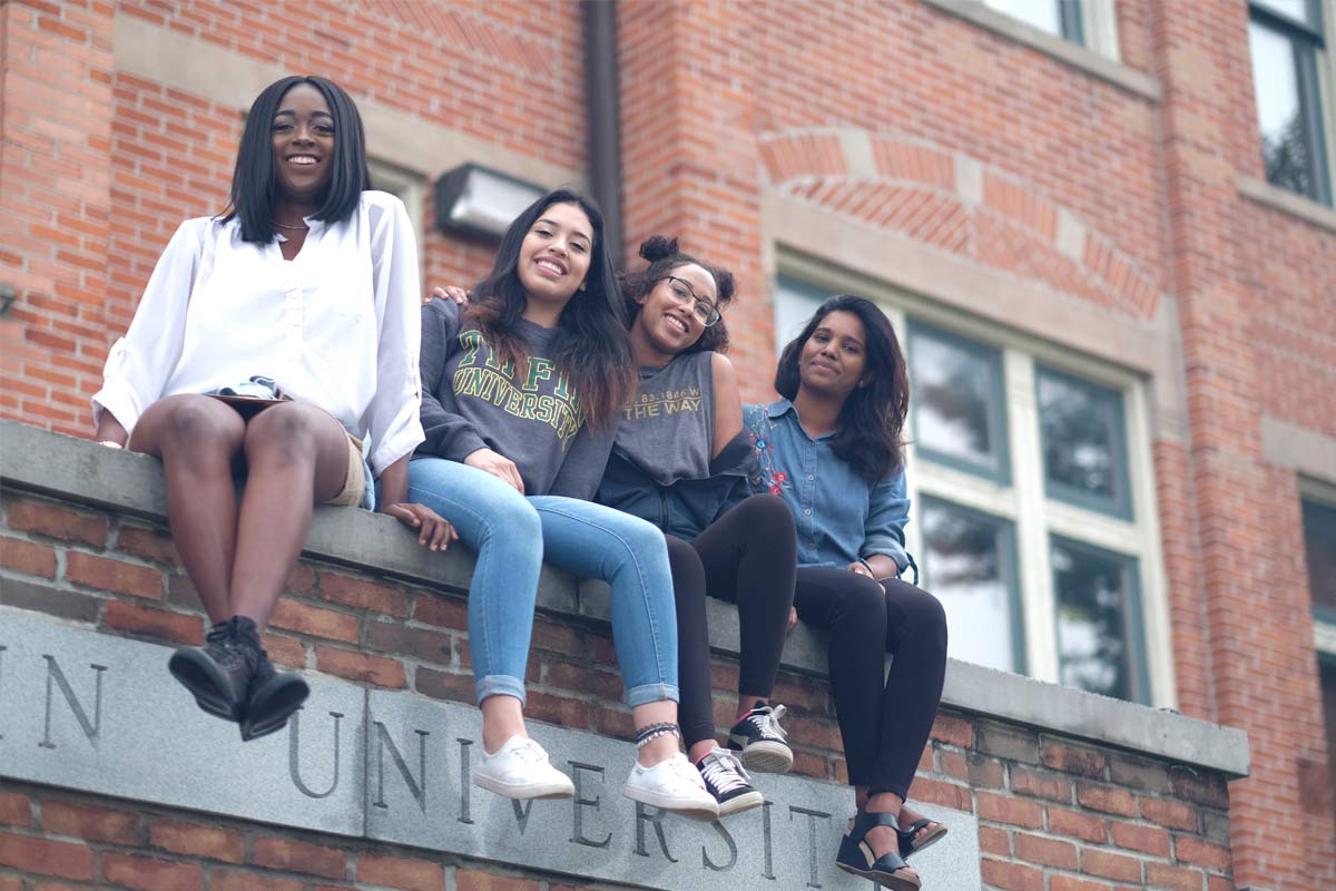 Four young women sitting on a brick ledge outside a university building, smiling at the camera.
