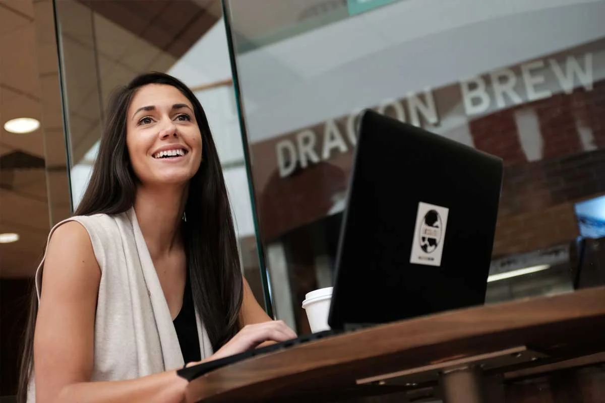 A woman with long dark hair smiling while sitting at a table with a laptop and coffee in a coffee shop.