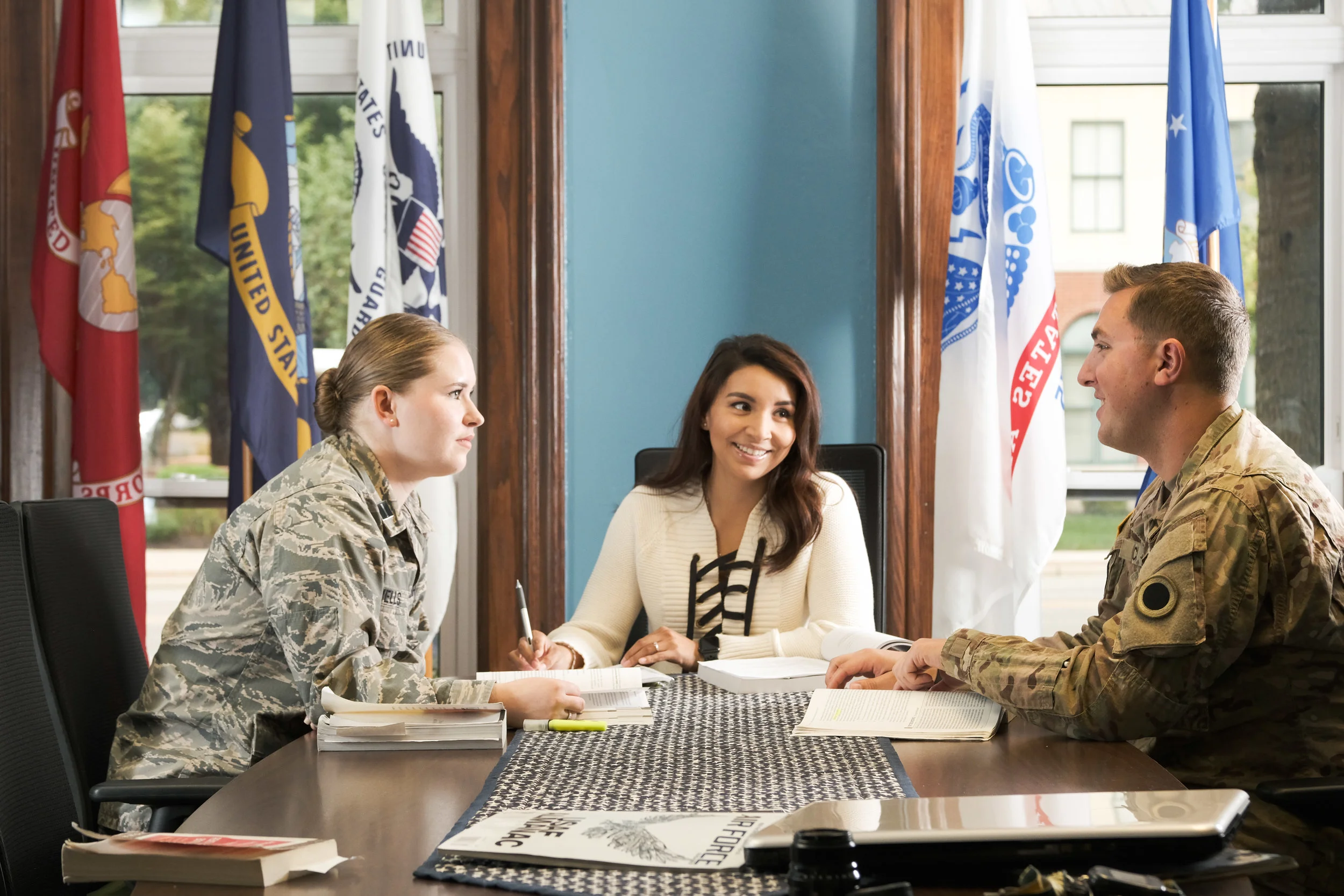 Three people, two in military uniforms and one woman in civilian clothing, having a discussion at a table in a conference room with flags in the background.