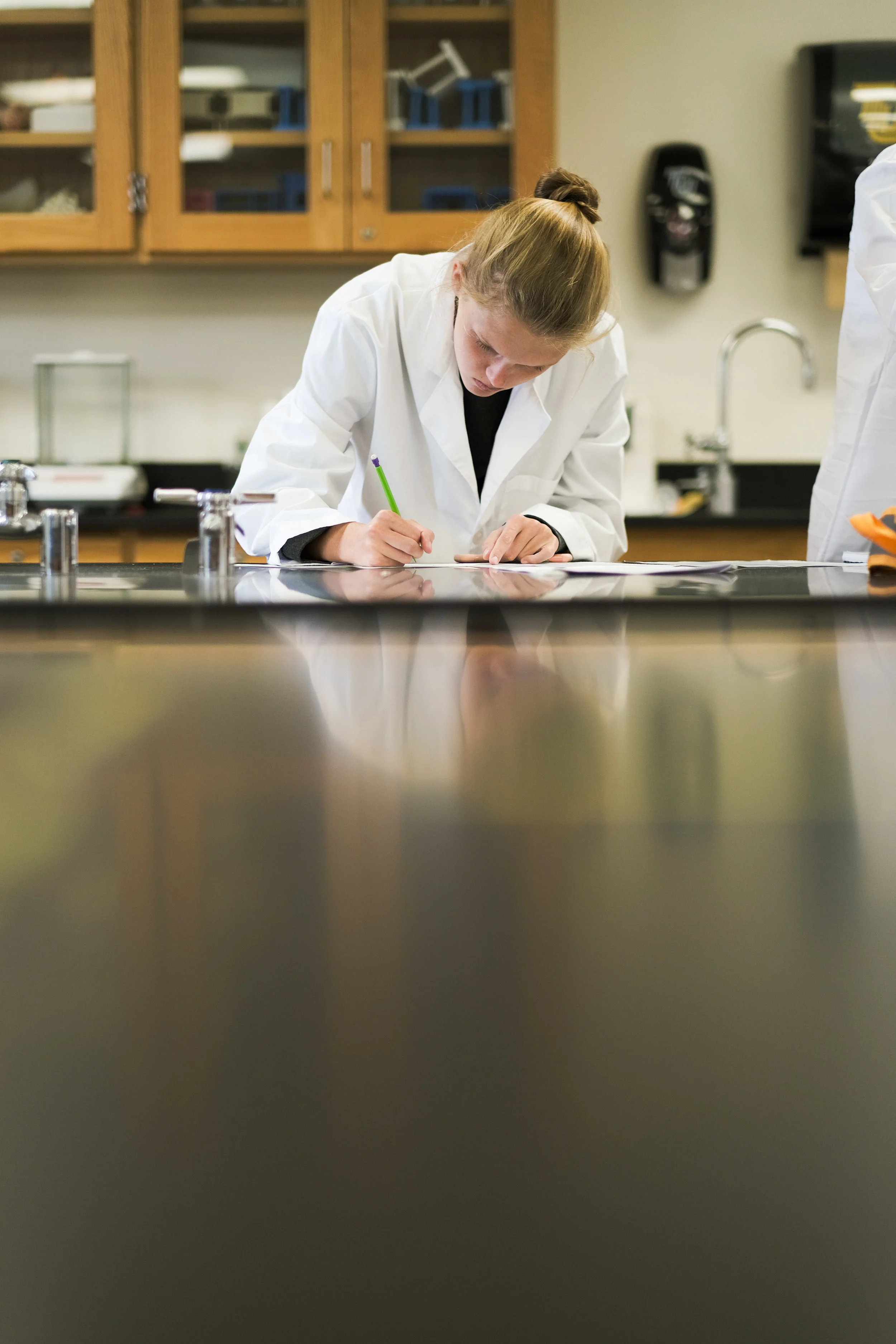 A young woman in a white lab coat writing on paper in a science classroom or laboratory.