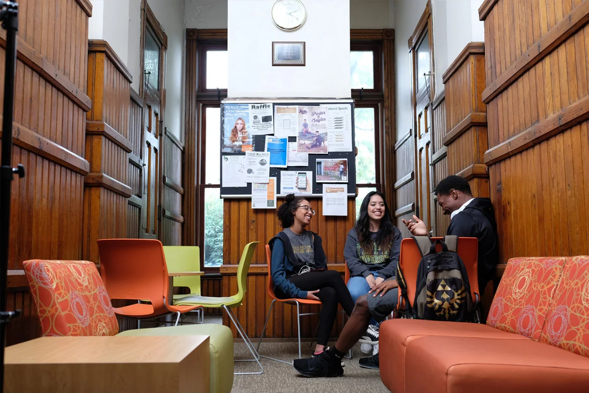 Three students sitting and chatting in a room with wooden walls and colorful chairs.