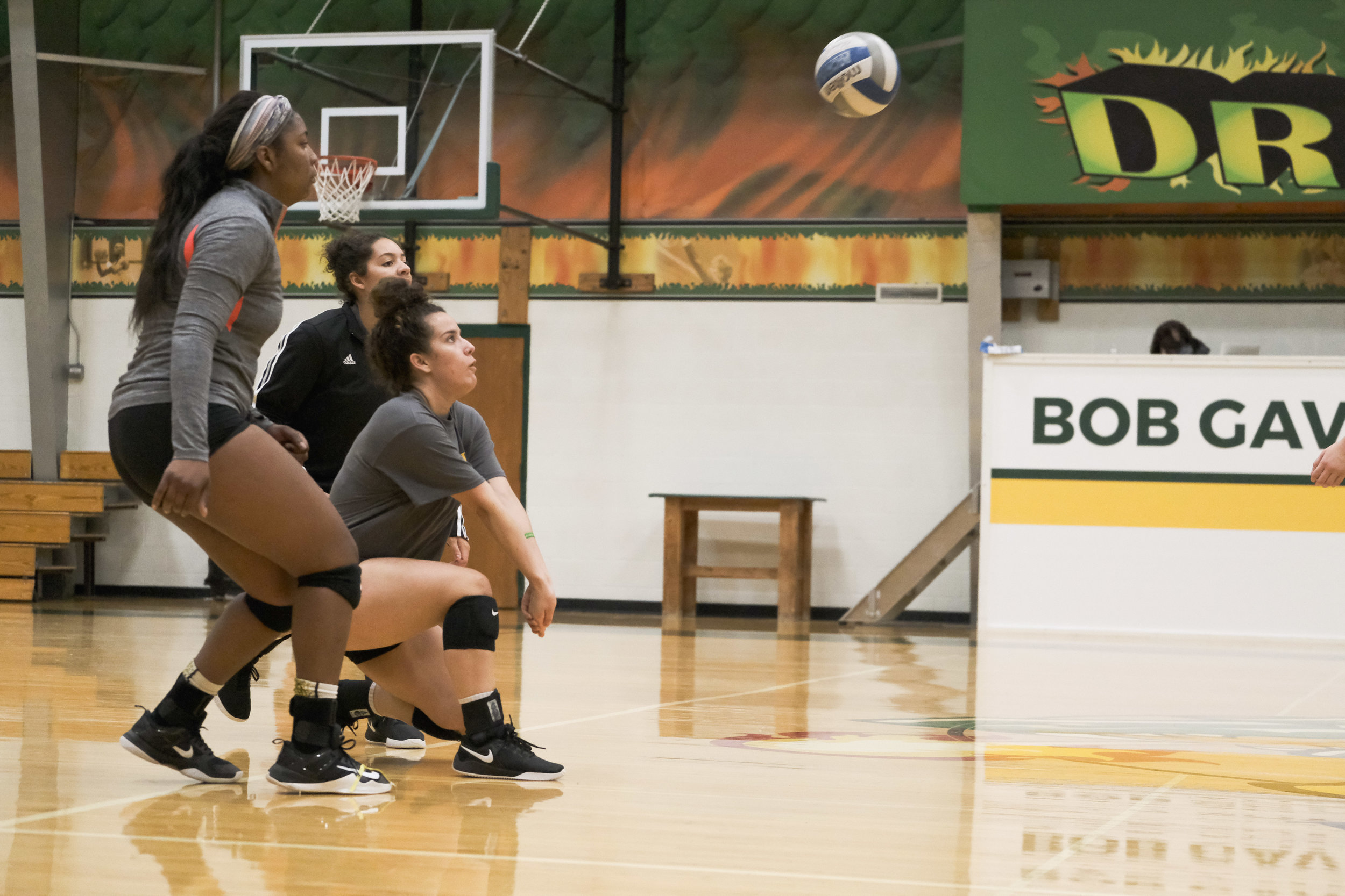 Three women playing volleyball in an indoor gym, one in a crouch ready to receive the ball, another standing, and the third squatting, all focused on the ball in the air.