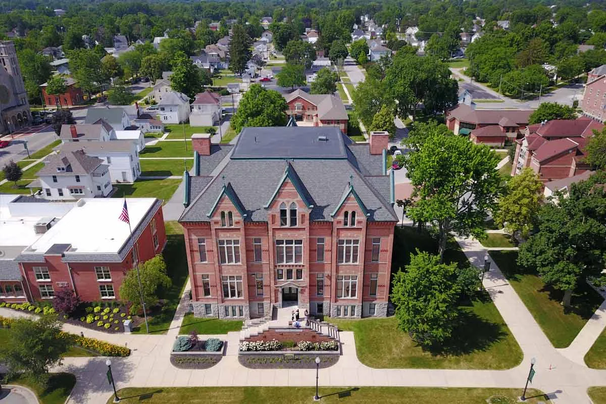 Aerial view of a historic red brick courthouse with a steep roof and large windows, surrounded by green trees and walkways in a small town.