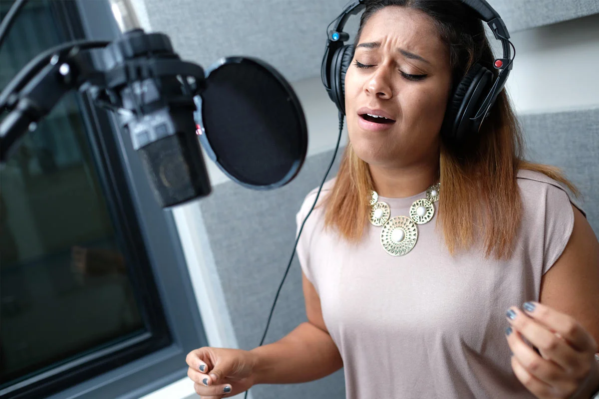 A woman with long brown hair wearing headphones and a beige top, singing passionately in a recording studio with a microphone and a pop filter.