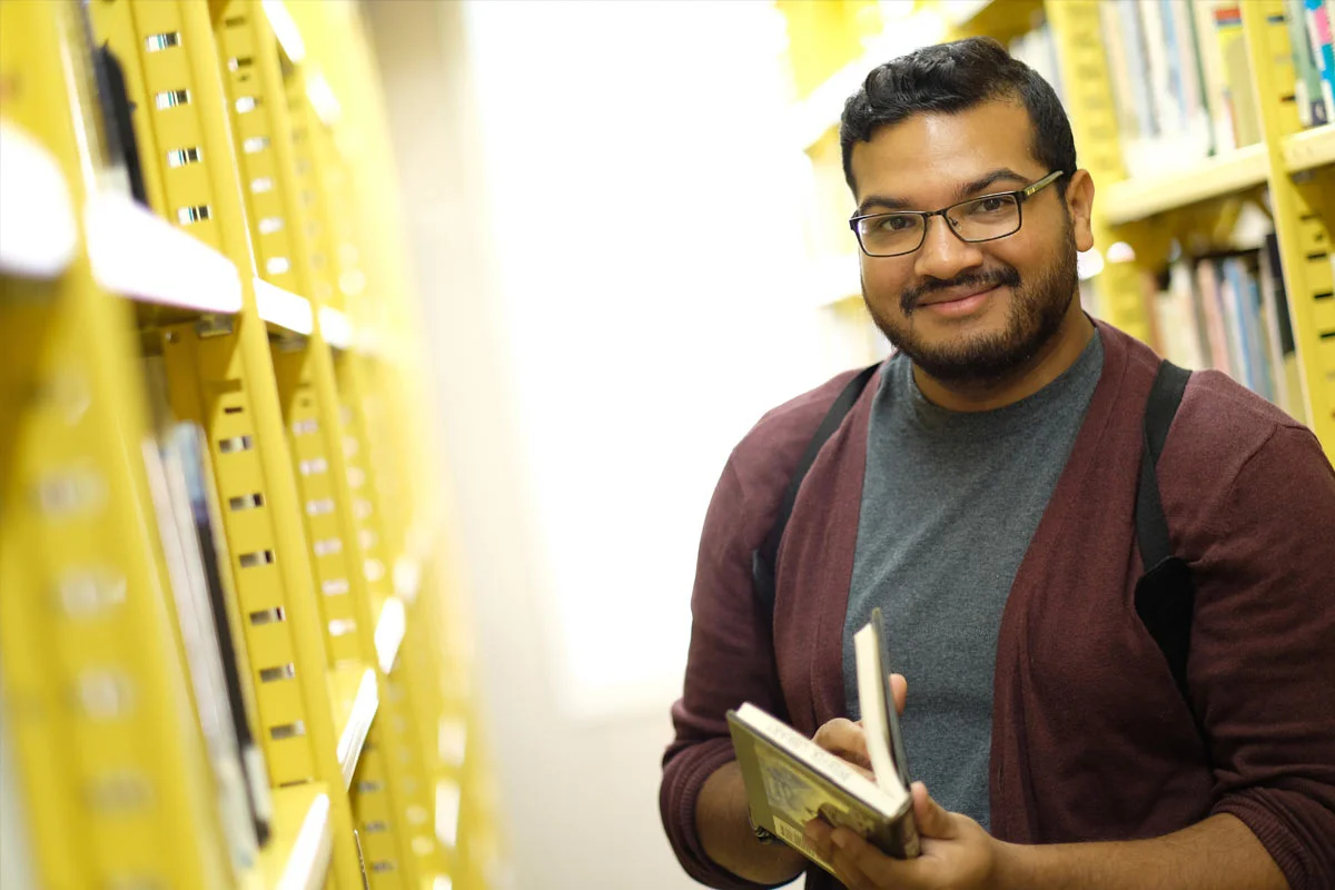 A young man with glasses and a beard standing in a library aisle, holding open a book and smiling at the camera.