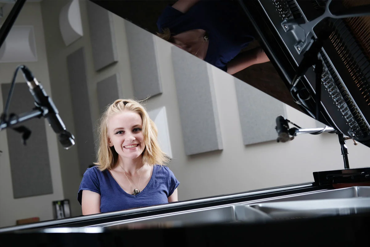 A young woman with blonde hair smiling at a grand piano in a music studio with soundproofing panels.