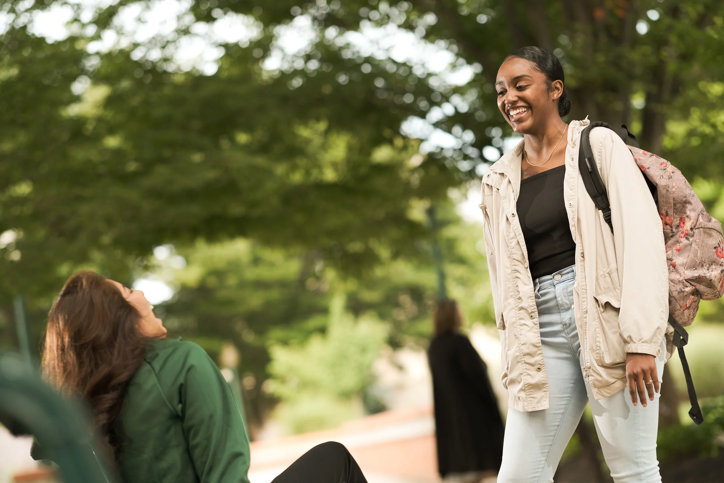 Two women conversing outdoors in a park, one sitting on a bench, the other standing with a backpack, surrounded by green trees.