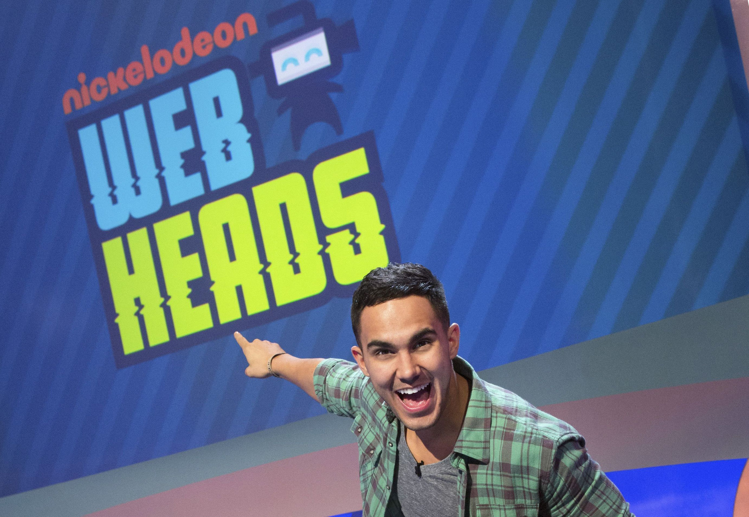 A young man smiling and pointing to a large blue sign that reads 'Nickelodeon UBER WINNER HERE' in bright colors.