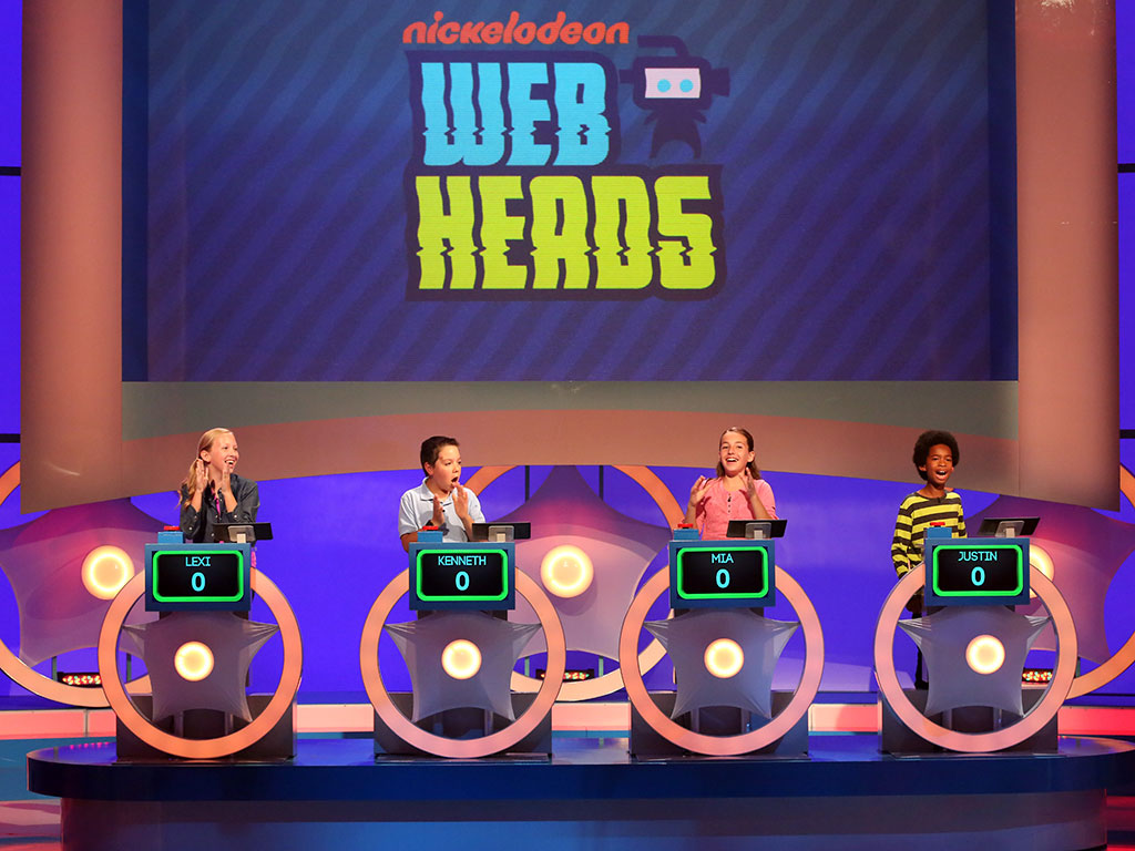 Children participating in a game show called 'Nickelodeon Web Heads,' sitting at individual podiums with digital displays. The large screen behind them shows the game's logo.