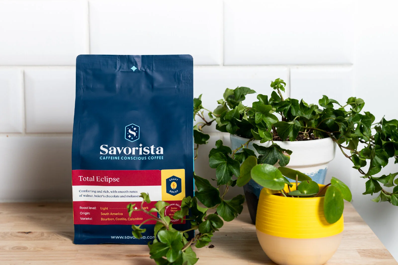 A bag of Savorista Total Eclipse coffee next to two potted plants, one in a white pot with greenery and another in a yellow and white striped pot. The background is a white wall with tiles.