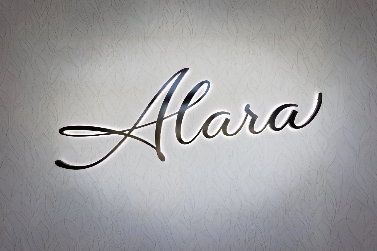 Illuminated nameplate with the word 'Alaia' in cursive script on a textured wall background.