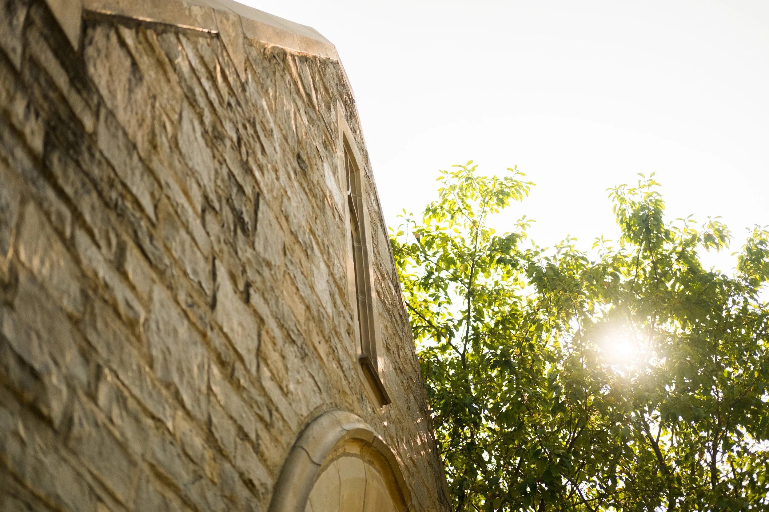 Close-up of a stone building with a window at an angle, sunlight shining through the trees on a bright day.