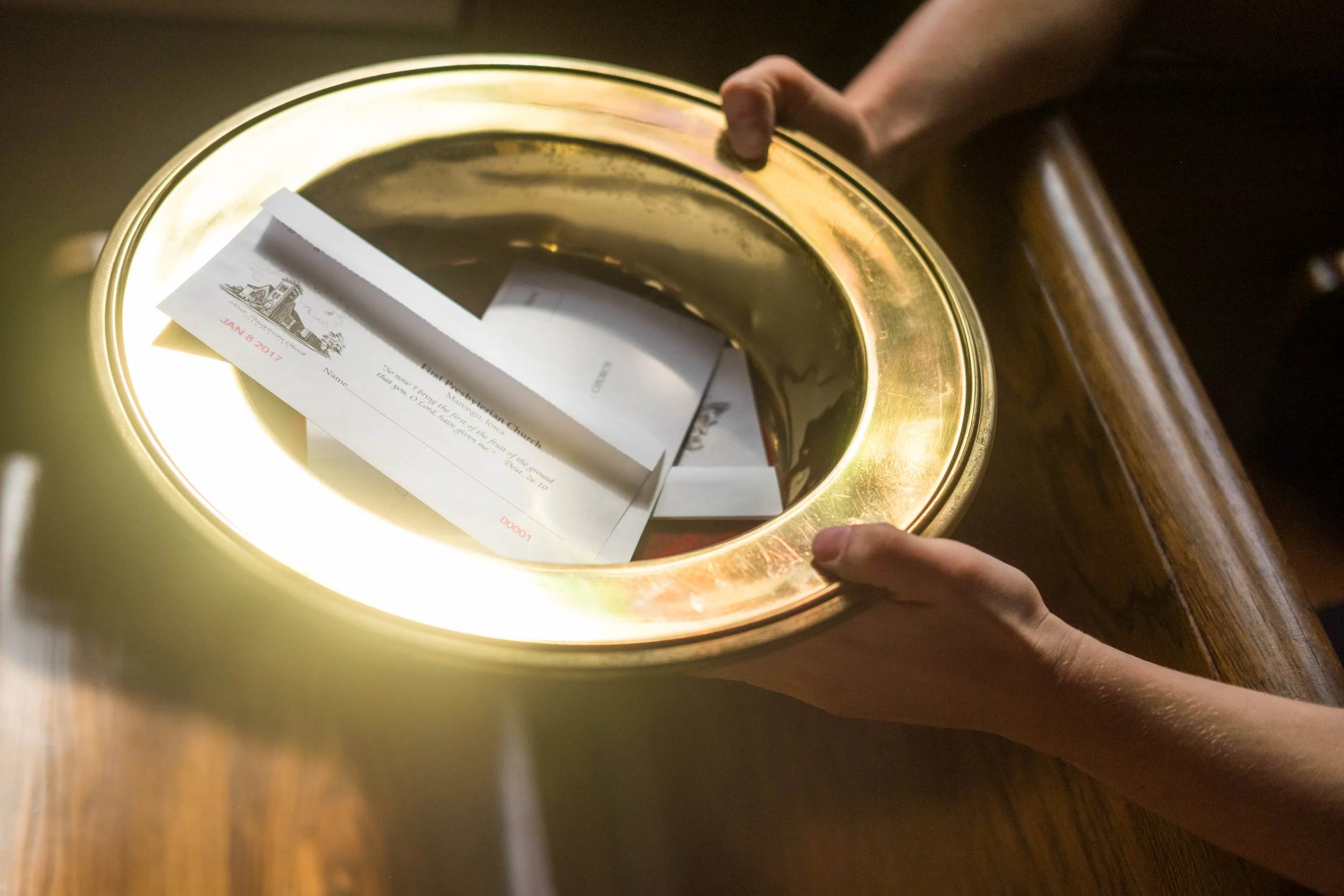 Person placing a donation envelope into a brass offering box in a church setting.