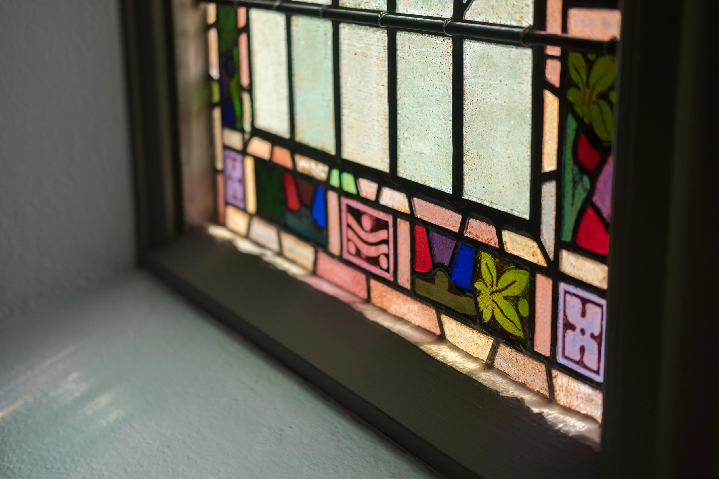 Close-up of a stained glass window with geometric and floral patterns, illuminated by sunlight, set in a wooden frame.