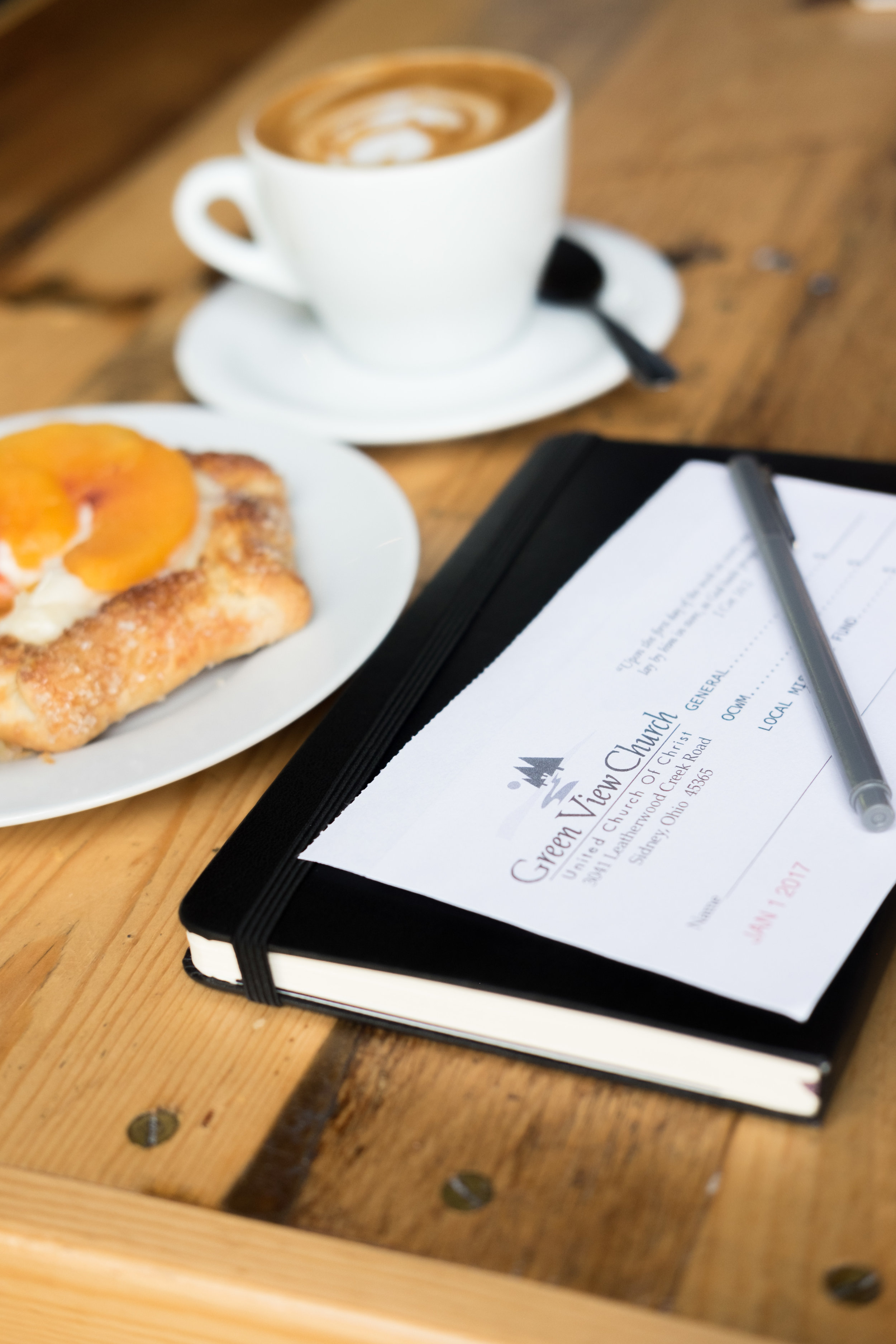 A wooden table with a breakfast plate, a coffee cup with a saucer, a spoon, and a black notebook with a pen resting on top. The notebook is labeled 'Green View Church' and has a date of January 2017.