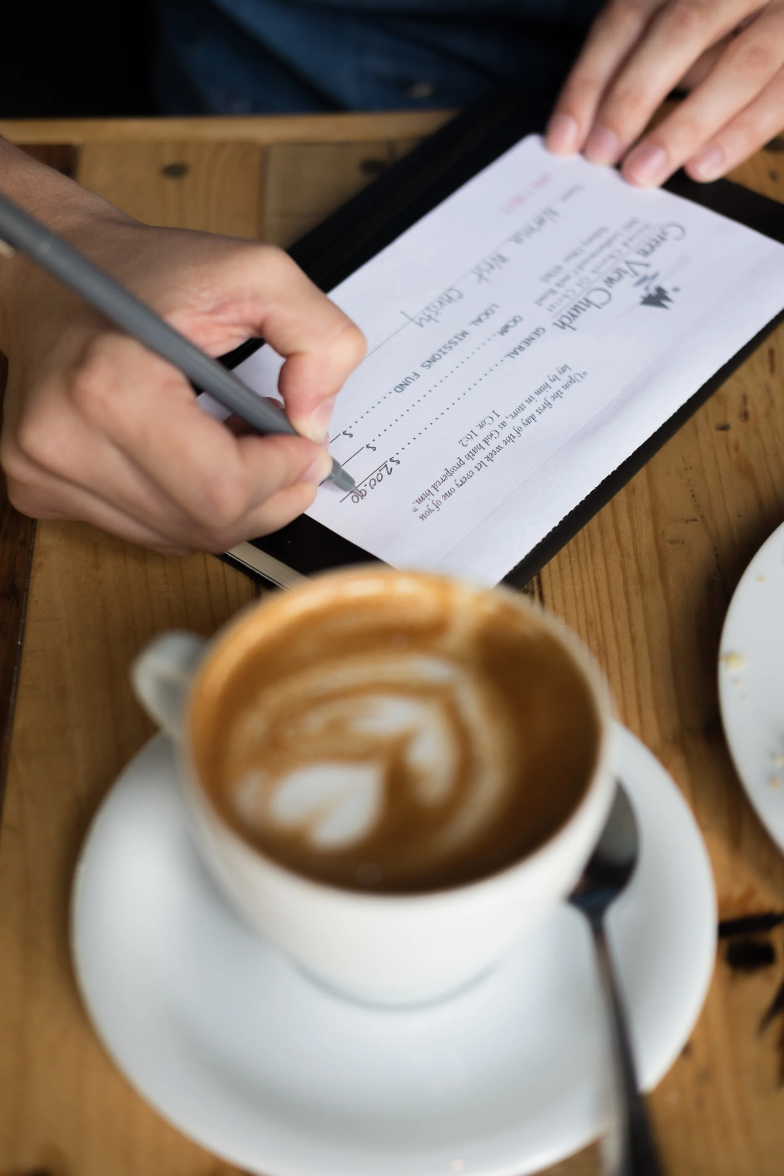 Person signing a check at a wooden table with a cup of coffee in the foreground.