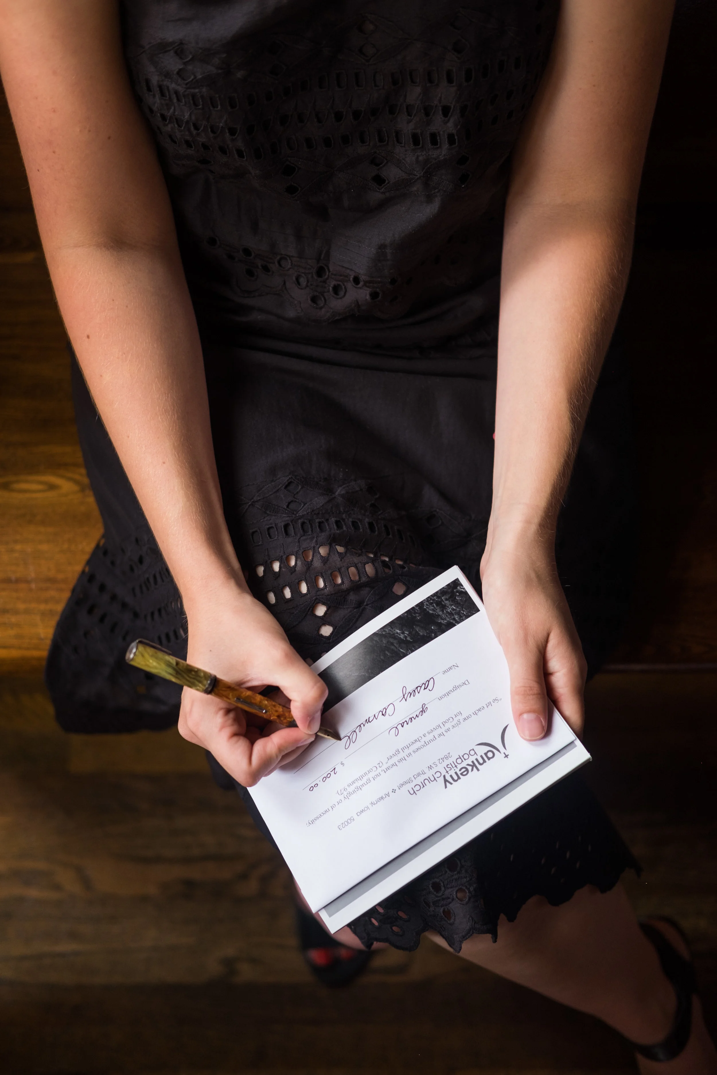 A person wearing a black dress with eyelet details, sitting on a wooden floor, signing a certificate with a gold-colored pen.